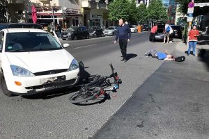 Ein Fahrrad liegt unter einem Auto. Der Fahrzeuglenker hat als Linksabbieger einen Fahrradfahrer übersehen. Dieser liegt aut der Straße und wartet auf die Feuerwehr.