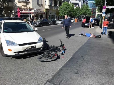Ein Fahrrad liegt unter einem Auto. Der Fahrzeuglenker hat als Linksabbieger einen Fahrradfahrer übersehen. Dieser liegt aut der Straße und wartet auf die Feuerwehr.
